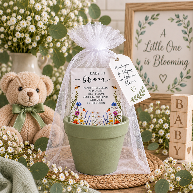A soft green terracotta pot holding a Baby in Bloom wildflower seed packet, wrapped in white organza with a thank-you tag, styled with a teddy bear, BABY blocks, daisies and a framed A Little One is Blooming sign