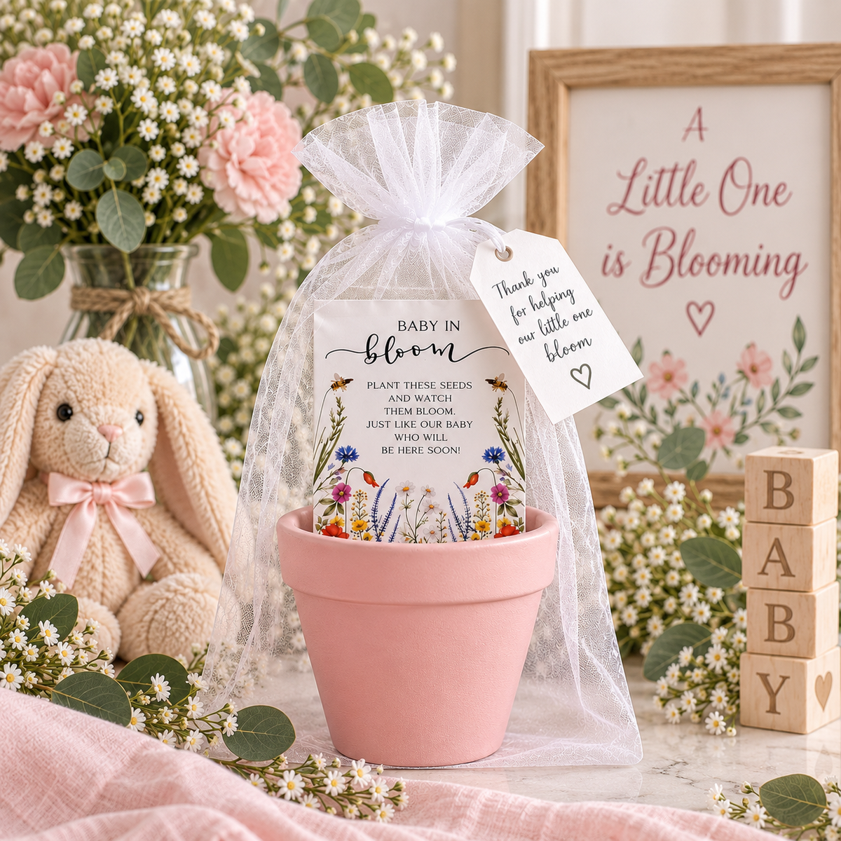 A pink terracotta pot holding a Baby in Bloom wildflower seed packet, wrapped in white organza with a thank-you tag, styled with a plush bunny, BABY blocks, pink carnations and a framed A Little One is Blooming sign
