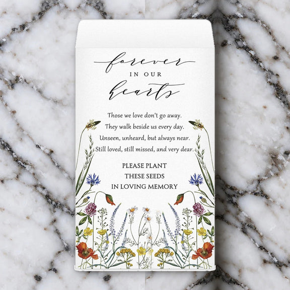 The Forever in Our Hearts memorial seed packet, printed with watercolour wildflowers and a remembrance verse, lying on white marble