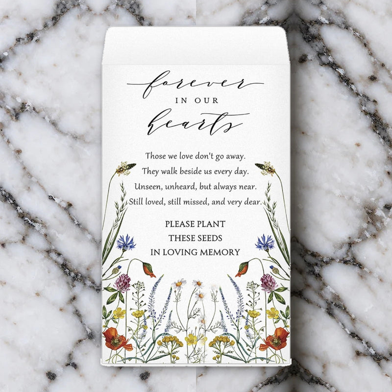 The Forever in Our Hearts memorial seed packet, printed with watercolour wildflowers and a remembrance verse, lying on white marble